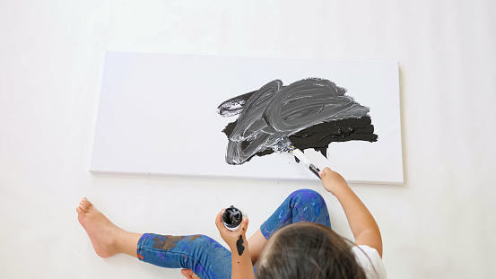 active little girl with space bun draws on sheet of paper with black paint and paintbrush sitting on atelier white floor close upper view