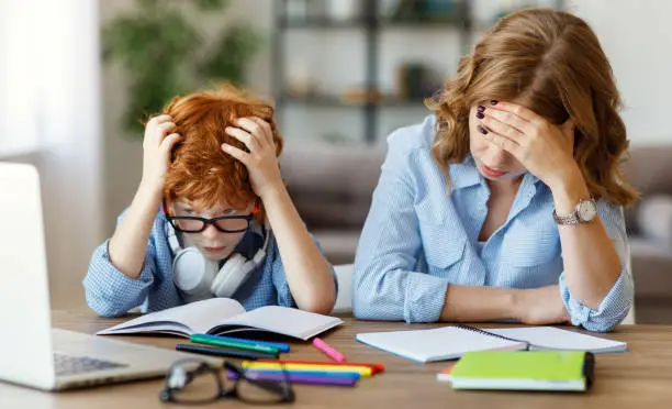 Stressed young woman grab head while sitting with son schoolboy at table with textbooks and notebook and studying difficult online homework