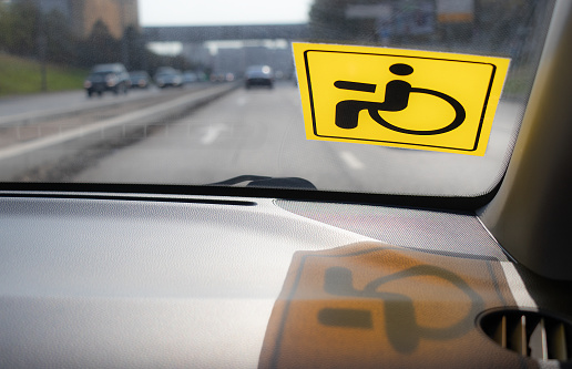 Yellow disabled driver sign on windshield of car. View from inside vehicle. Reflection of sticker on dashboard. Blurred silhouettes of cars and highway on background