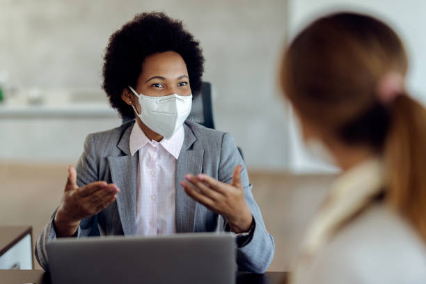 Black female financial consultant talking to her client and wearing protective face mask during the meeting. African American bank manager with protective face mask communicating with her client during a meeting in the office. business meeting 2 people stock pictures, royalty-free photos & images
