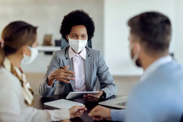 Photo of Happy African American financial advisor consulting her clients while wearing protective face mask on a meeting.