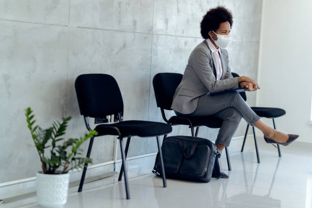 Black pensive businesswoman with face mask waiting for job interview. Distraught African American businesswoman wearing protective face mask while sitting in a hallway and waiting for job interview. job search stock pictures, royalty-free photos & images