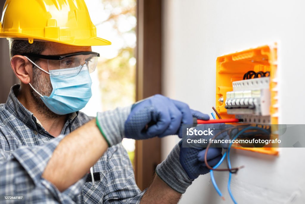 Electrician at work with face protected by surgical mask to prevent Coronavirus infection. Covid-19 pandemic. Electrician at work on an electrical panel protected by helmet, safety goggles and gloves; wear the surgical mask to prevent the spread of Coronavirus. Construction industry. Covid-19 Prevention. Electrician Stock Photo
