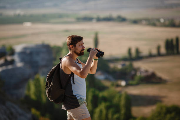 un tipo mirando binoculares en la colina. hombre en camiseta con mochila - pursuit binoculars mountain sky fotografías e imágenes de stock