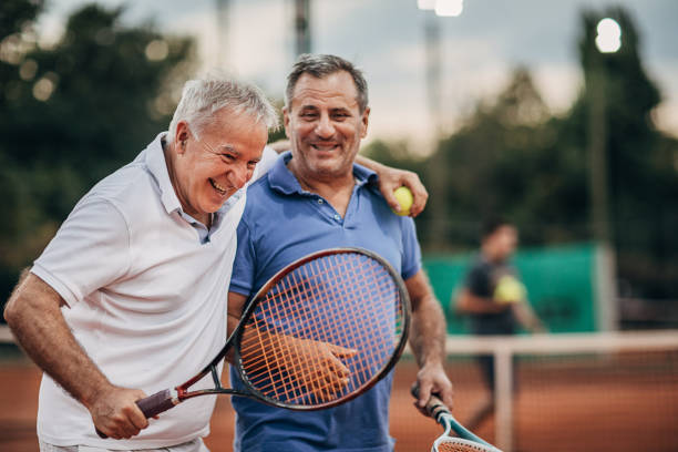 Two cheerful senior men talking while walking on the outdoor tennis court Two cheerful senior tennis players leaving tennis court after a match while smiling and hugging. Concept of healthy and fit senior people. racket sport stock pictures, royalty-free photos & images