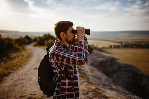 un tipo mirando binoculares en la colina. hombre en camiseta con mochila - pursuit binoculars mountain sky fotografías e imágenes de stock