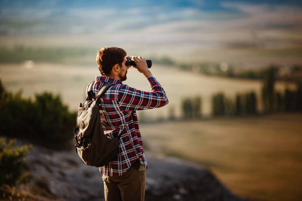 언덕에서 쌍안경을 보고 있는 남자. 배낭을 가진 티셔츠를 입은 남자 - pursuit binoculars mountain sky 뉴스 사진 이미지