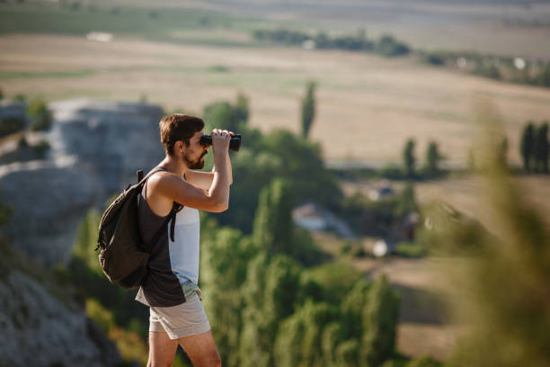 언덕에서 쌍안경을 보고 있는 남자. 배낭을 가진 티셔츠를 입은 남자 - pursuit binoculars mountain sky 뉴스 사진 이미지