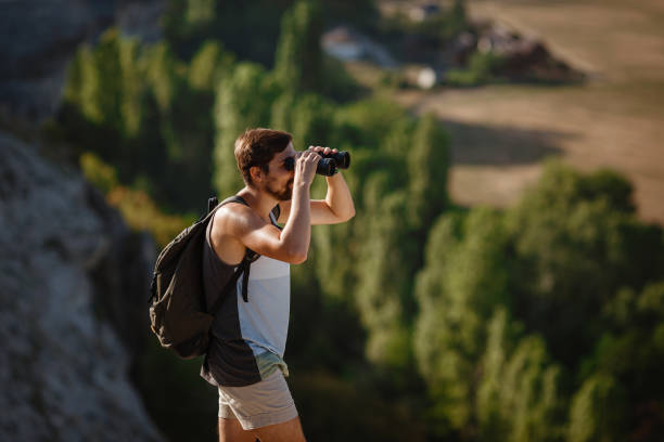 un tipo mirando binoculares en la colina. hombre en camiseta con mochila - pursuit binoculars mountain sky fotografías e imágenes de stock