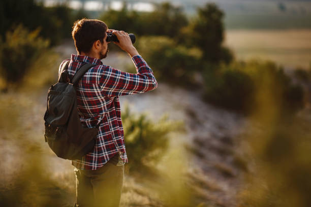 un tipo mirando binoculares en la colina. hombre en camiseta con mochila - pursuit binoculars mountain sky fotografías e imágenes de stock