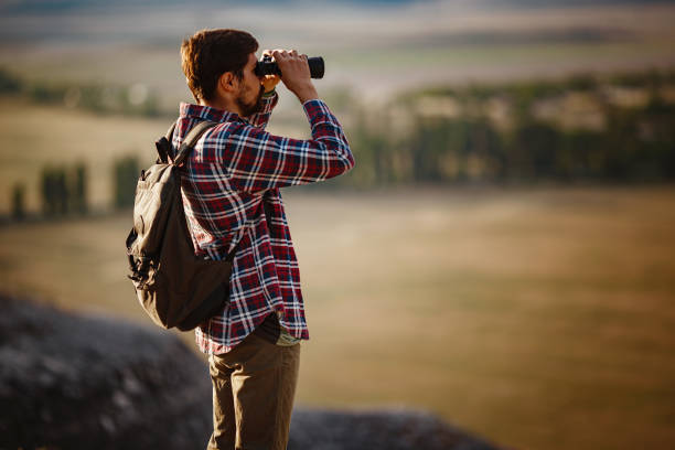 un tipo mirando binoculares en la colina. hombre en camiseta con mochila - pursuit binoculars mountain sky fotografías e imágenes de stock