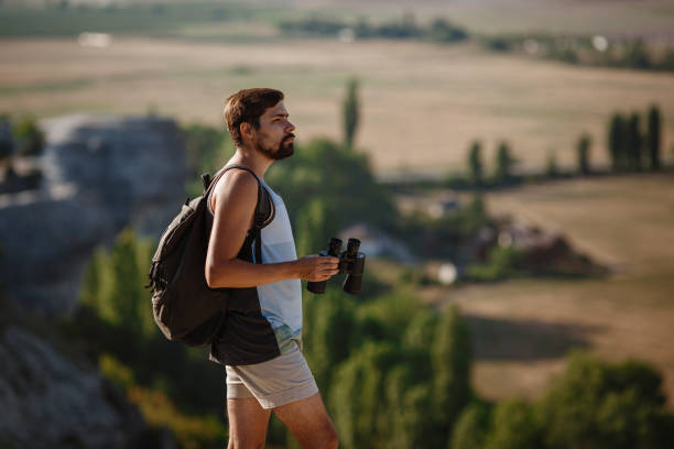 un tipo mirando binoculares en la colina. hombre en camiseta con mochila - pursuit binoculars mountain sky fotografías e imágenes de stock