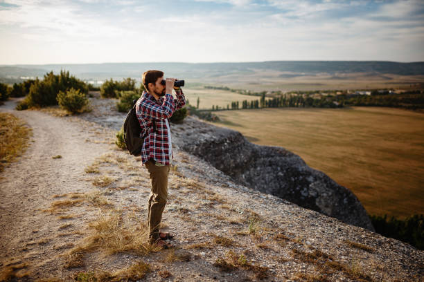 un tipo mirando binoculares en la colina. hombre en camiseta con mochila - pursuit binoculars mountain sky fotografías e imágenes de stock