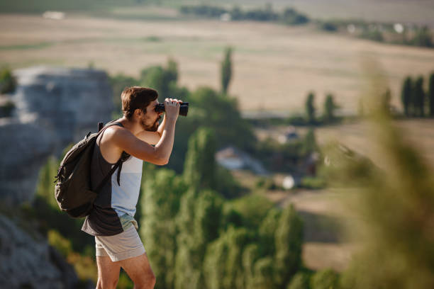 언덕에서 쌍안경을 보고 있는 남자. 배낭을 가진 티셔츠를 입은 남자 - pursuit binoculars mountain sky 뉴스 사진 이미지