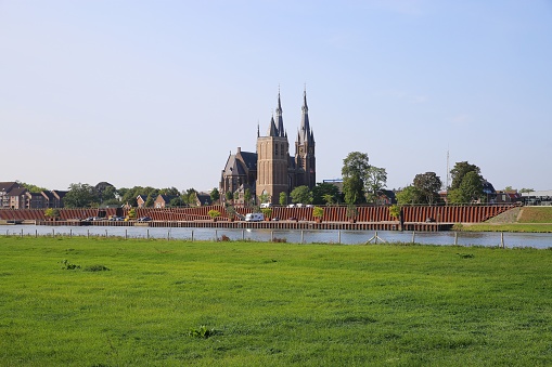 Cuijk, Netherlands - August 9. 2020: View over green pasture and river maas on cityscape of dutch village with cathedral (focus on church)