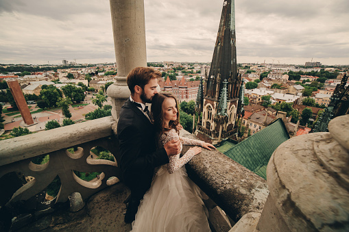 Happy newlywed. beautiful bride and stylish groom are hugging on the balcony of old gothic cathedral with panoramic city views.