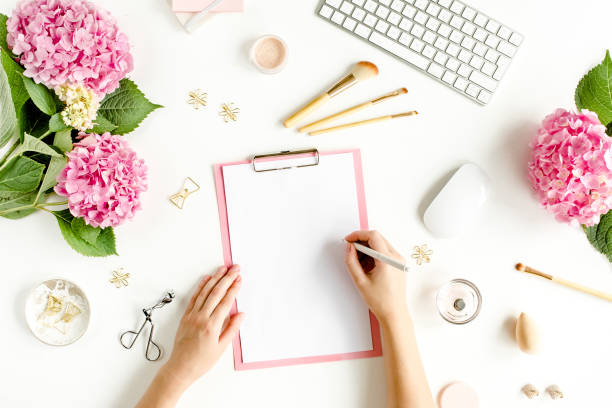 istock Female workspace with female hands, clipboard, bouquet hydrangea, computer, accessories on white background. Stylized women's desk. Flat lay. Top view 1272477031