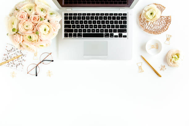 istock Stylized women's home office desk. Workspace with laptop, computer, bouquet ranunculus and roses, clipboard on white background. Flat lay. Top view 1272478922