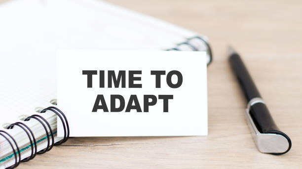 istock TIME TO ADAPT on a card next to a notebook with a pen on the table. Business concept 1272480010