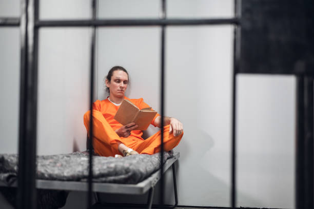 A young inmate with a tattooed face is being remotely trained in a cell. Reads a textbook sitting on a bunk stock photo