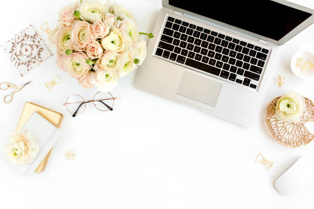 istock Stylized women's home office desk. Workspace with laptop, computer, bouquet ranunculus and roses, clipboard on white background. Flat lay. Top view 1272484728