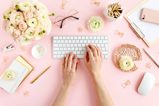 istock Stylized, pink women's home office desk. Workspace with female hands, computer, bouquet ranunculus and roses, clipboard on pink background. Flat lay 1272486369