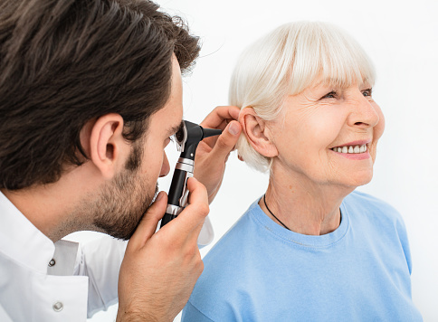 ENT doctor examining senior patient ear, using an otoscope, in doctors office. Elderly smiling woman getting medical ear exam at clinic