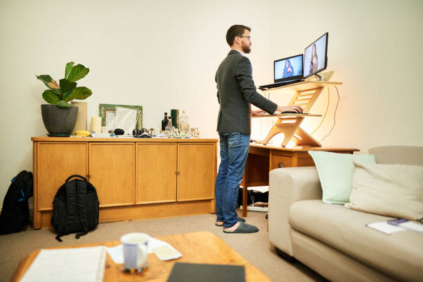 Young man working remotely at a standing desk in his living room Young man having a online video meeting with office colleagues at a standing desk while working remotely from home ergonomics photos stock pictures, royalty-free photos & images