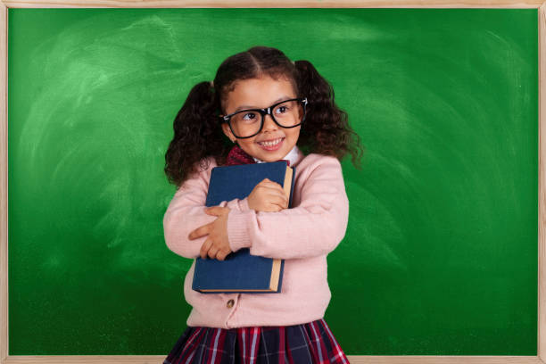 istock Little girl with glasses reading a book in front blackboard 1272494519