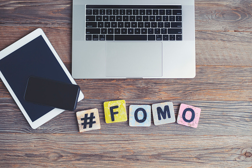 FOMO, Fear OF Missing Out. Laptop, digital tablet and mobile phone with a hashtag and the letters FOMO beside them. They are on a wooden background with copy space.