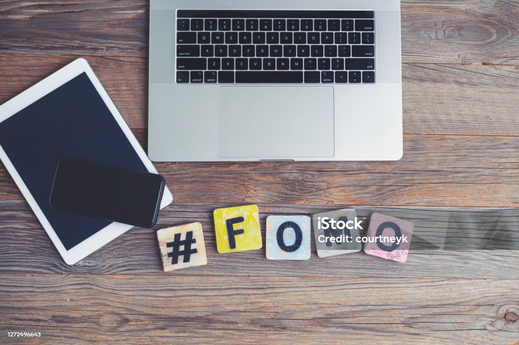FOMO, Fear OF Missing Out. Laptop, digital tablet and mobile phone with a hashtag and the letters FOMO beside them. FOMO, Fear OF Missing Out. Laptop, digital tablet and mobile phone with a hashtag and the letters FOMO beside them. They are on a wooden background with copy space. Acronym Stock Photo