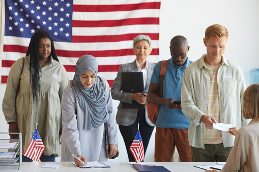 Multi-ethnic group of people registering at polling station decorated with American flags on election day, focus on Arab woman signing ballot form in foreground, copy space