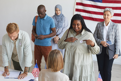 Multi-ethnic group of people registering at polling station decorated with American flags on election day, focus on smiling African woman pointing at I VOTE sticker, copy space