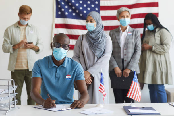 People Wearing Masks at Voting Station Multi-ethnic group of people standing in row and wearing masks at polling station on election day, focus on African-American man registering for voting ballot measure stock pictures, royalty-free photos & images