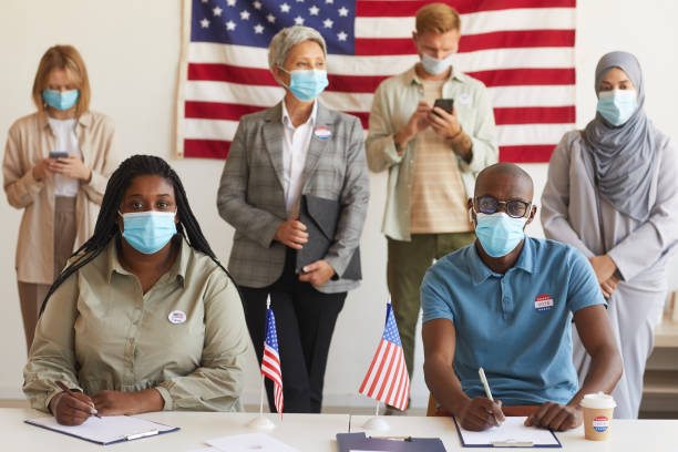 Group of People Voting at Polling Station Multi-ethnic group of people standing in row and wearing masks at polling station on election day, focus on African-American couple looking at camera while registering for voting ballot measure stock pictures, royalty-free photos & images