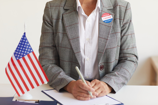 Cropped portrait of mature woman sitting at desk with American flag at polling station on election day and registering for voting