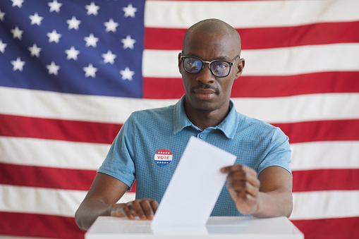 Portrait of modern African-American man putting vote bulletin in ballot box and looking at camera while standing against American flag on election day, copy space