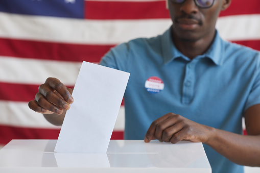 Cropped portrait of African-American man putting vote bulletin in ballot box while standing against American flag on election day, copy space