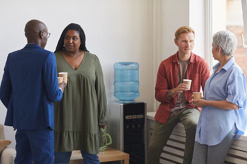 Multi-ethnic group of people drinking coffee and chatting while standing by water cooler in support meeting, copy space