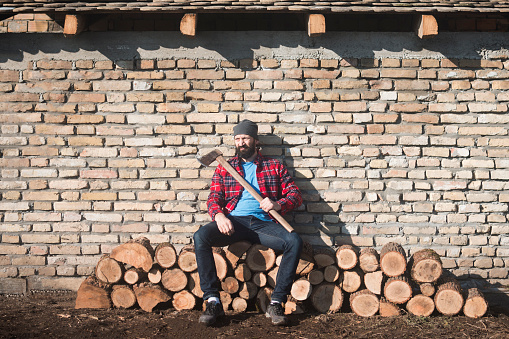 The lumberjack is sitting with his axe on tree trunks on brick wall background.