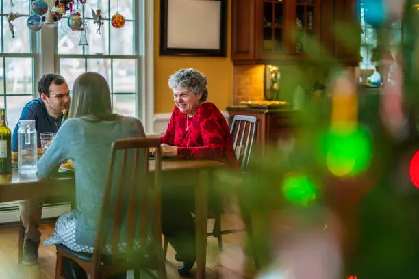 Photo of Two-generation family, the senior woman, a mother with adult children, gathered together and have the Christmas dinner. The scene is partially covered by the blurred Christmas Tree in the foreground.