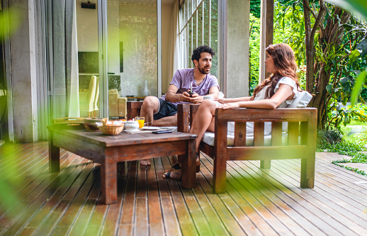 Low angle view of bearded mixed race man and long haired Hispanic woman sitting on outdoor home deck and enjoying breakfast and conversation.