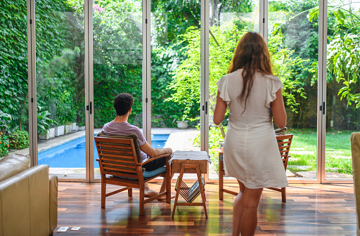 Young couple relaxing in open plan sitting area with naturally lit view to lush backyard and swimming pool.