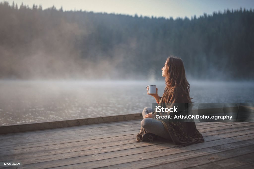 Woman Relaxing In Nature. Side view of woman sitting on wooden pier in nature, she looking the sunrise and drinking coffee. Relaxation Stock Photo