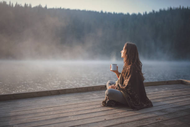 Woman Relaxing In Nature. Side view of woman sitting on wooden pier in nature, she looking the sunrise and drinking coffee. coffee drink stock pictures, royalty-free photos & images