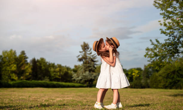 Side view of sisters in similar dresses hugging and kissing each other while resting in park on sunny summer day