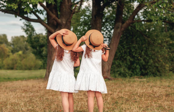 Back view of unrecognizable sisters in similar dresses and hats admiring tall tree on summer day in green park