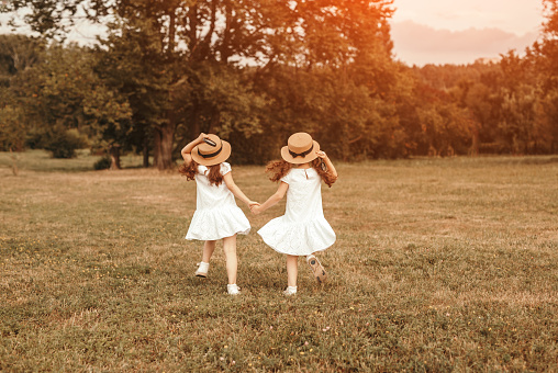 Back view of unrecognizable sisters in similar outfits holding hands and running on grass while playing in summer park together