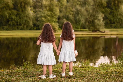 Back view of unrecognizable girls in similar white dresses holding hands and admiring calm lake while resting on grassy shore in summer park together