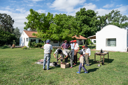 Argentine gaucho boys, teens, and adults relaxing outdoors while grilling meat and frying empanadas for midday asado meal.